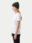 A woman in profile wearing a white short-sleeve crewneck t-shirt, black pants, and an olive green ribbed knit beanie, photographed against a light gray studio background.
