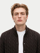 A young man with light brown hair wearing a dark brown cable-knit button-up cardigan over a white t-shirt, photographed against a solid light gray background.