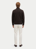 A man viewed from behind wearing a dark brown cable-knit sweater, off-white trousers, and dark brown loafers, set against a plain light gray studio background.