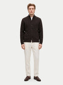 A man modeling a dark brown cable-knit cardigan with a stand collar, worn over a white shirt and paired with off-white trousers and brown leather loafers, against a light gray studio background.