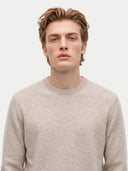 A man wearing a light beige crewneck knit sweater, photographed from the chest up against a plain light gray background.