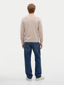 A man viewed from behind wearing a light beige crewneck sweater, blue jeans, and off-white sneakers, standing against a plain light gray studio background.