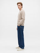 A man stands in profile wearing a light beige crewneck sweater, dark blue wide-leg jeans, and off-white sneakers against a plain white studio background.