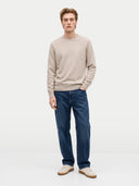 A man modeling a light beige crewneck sweater and dark blue jeans against a plain white studio background.