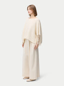 side view of a woman wearing a beige cashmere poncho with fringed sleeves, over a white outfit. #color_white