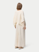 back view of a woman wearing a beige cashmere poncho with fringed sleeves, over a white outfit. #color_white