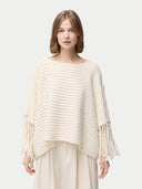 A woman wearing a beige cashmere poncho with fringed sleeves, over a white outfit. #color_white