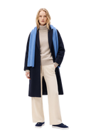 Woman in a black oval-sleeve cashmere long coat over a beige turtleneck sweater, with a light blue scarf, cream textured knit trousers and navy loafers