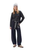 Woman in a charcoal rib-knit cashmere sweater and a light grey beanie, a rope-knot print shawl tied at the waist, charcoal barrel-leg knit trousers and beige loafers
