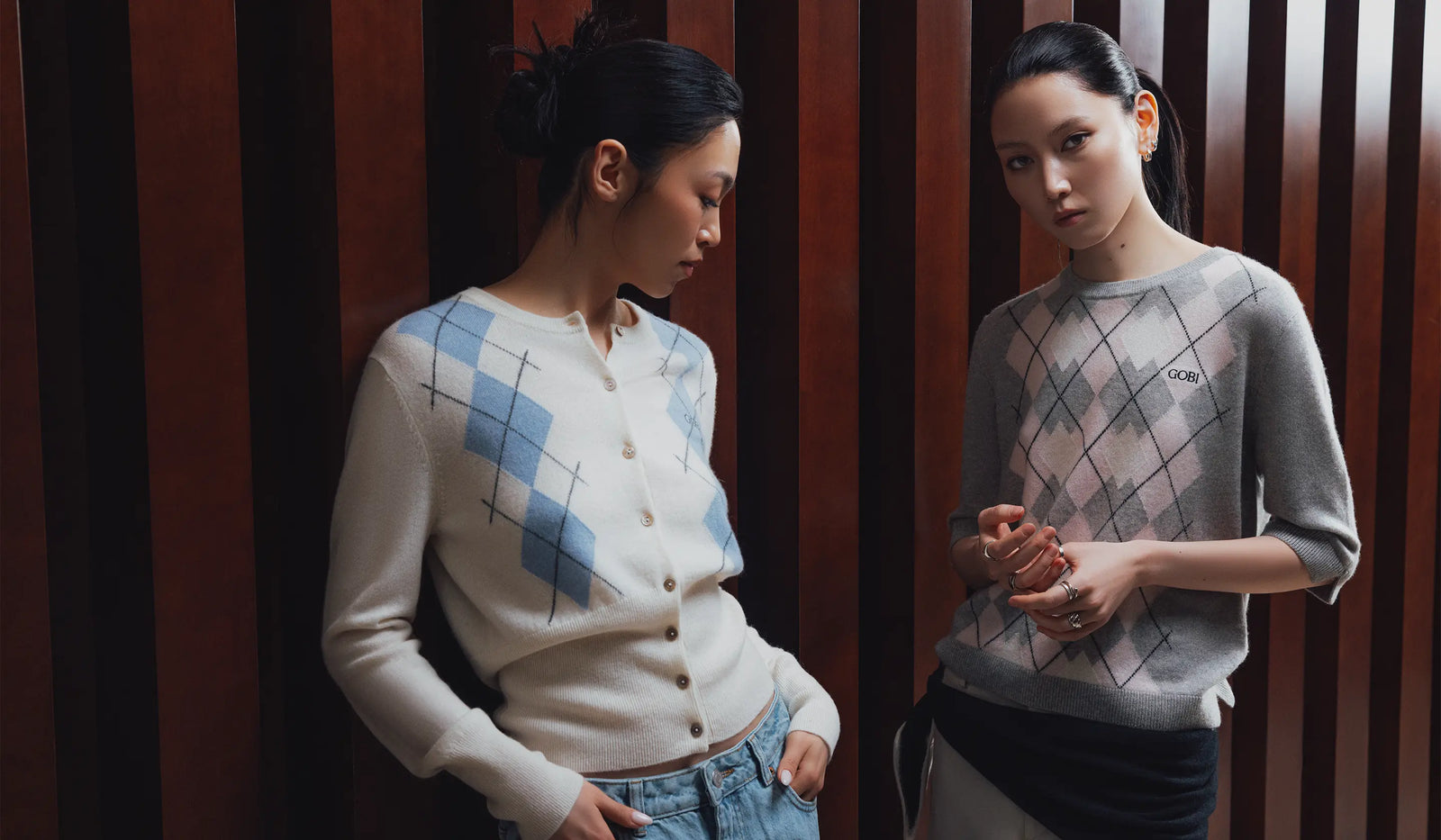 Two women wearing argyle pattern sweaters standing against a wooden paneled wall. desktop