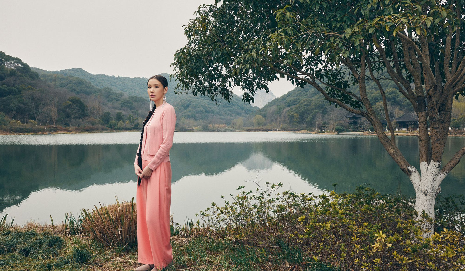 Woman in a pink dress standing by a lake with mountains in the background