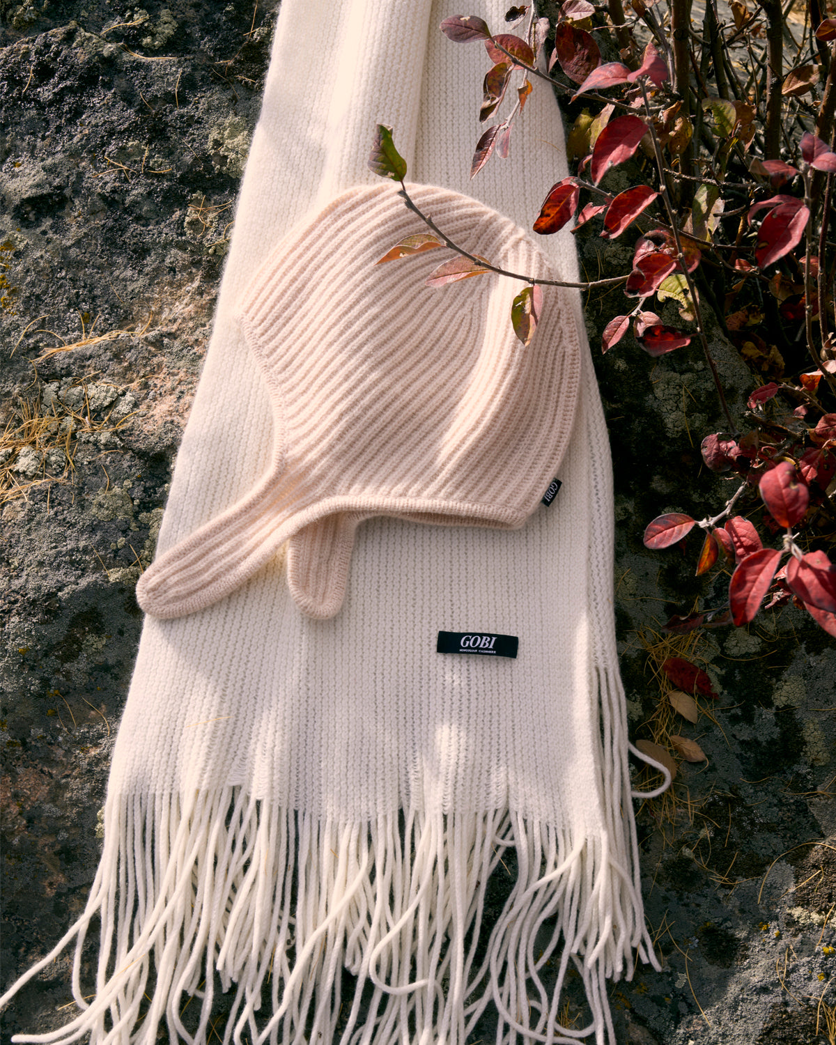 Cream ribbed cashmere scarf and trapper hat laid on a lichen-covered rock beside red autumn leaves