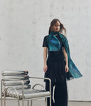 Woman wearing a blue scarf standing next to a metallic chair against a gray wall.