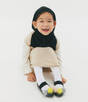 Child wearing a black headscarf, beige sweater, white tights, and black sandals with colorful pom-poms on a white background