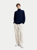 Man wearing a navy turtleneck sweater and white pants on a light gray background