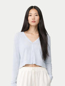 A woman with long dark hair wearing a light blue cropped V-neck cardigan and white pants, photographed against a plain light gray studio background. #color_soft-ice