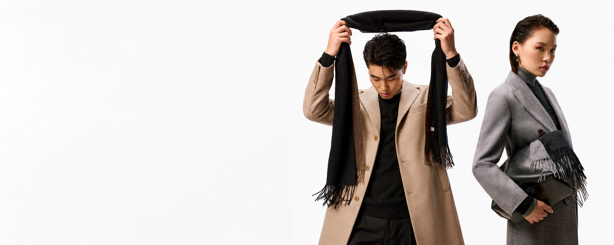A man in a taupe cashmere coat lifts a black fringed scarf above his head, beside a woman in a gray coat holding a black leather bag and a fringed scarf