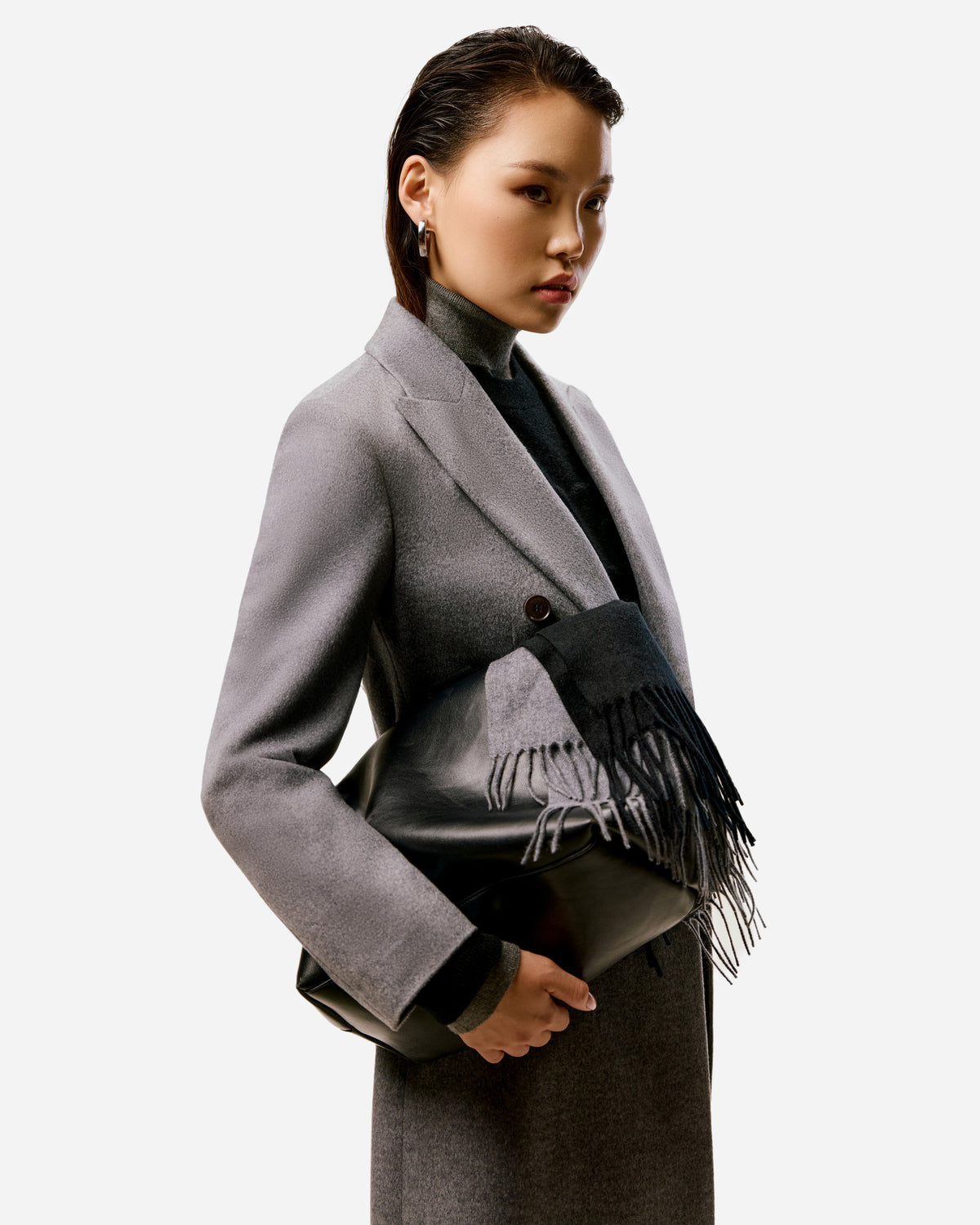 Woman in a gray cashmere coat holding a black leather bag draped with a gray and black fringed scarf