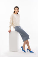 A woman in a cream cashmere sweater and grey skirt seated on a white block