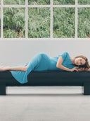 Woman in a blue dress lying on a black sofa with greenery outside the window. mobile