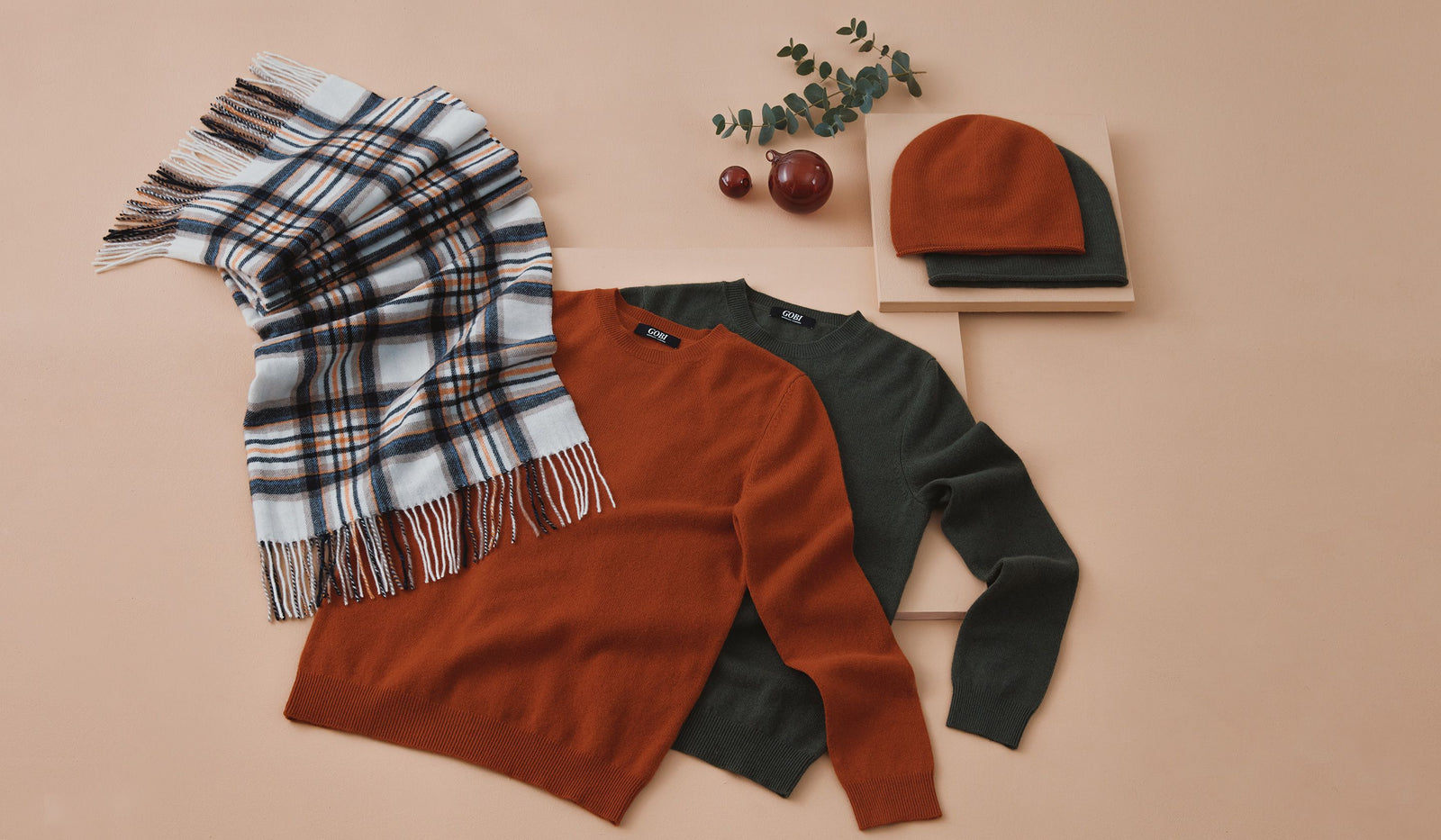 Plaid scarf, brown sweater, green sweater, and red beanie on a beige background. desktop