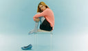 Woman sitting on a chair with one leg crossed over the other, wearing a pink sweater and black pants, on a light blue background.  desktop