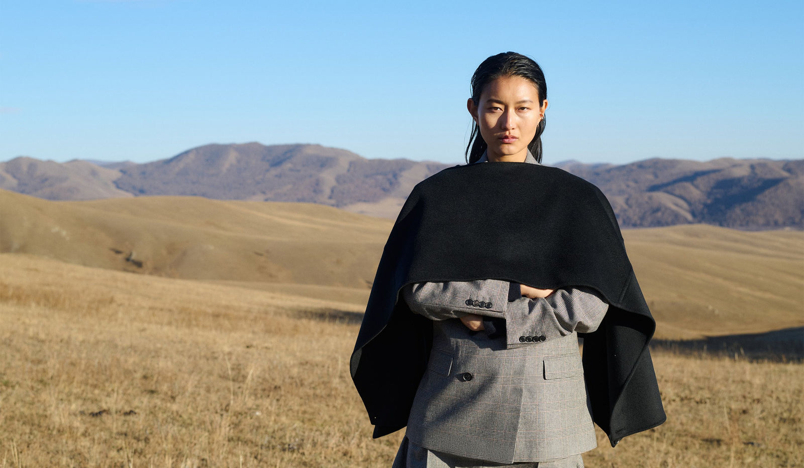 Person wearing a black cape over a gray outfit in a desert landscape with mountains in the background.  desktop