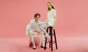 Two people sitting on a white chair and black stool against a pink background.  desktop
