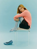 Woman sitting on a clear stool against a light blue background, wearing a pink sweater and gray shoes. mobile