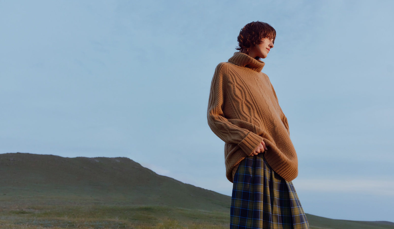 Person wearing a brown sweater and plaid skirt standing in a field with mountains in the background. desktop