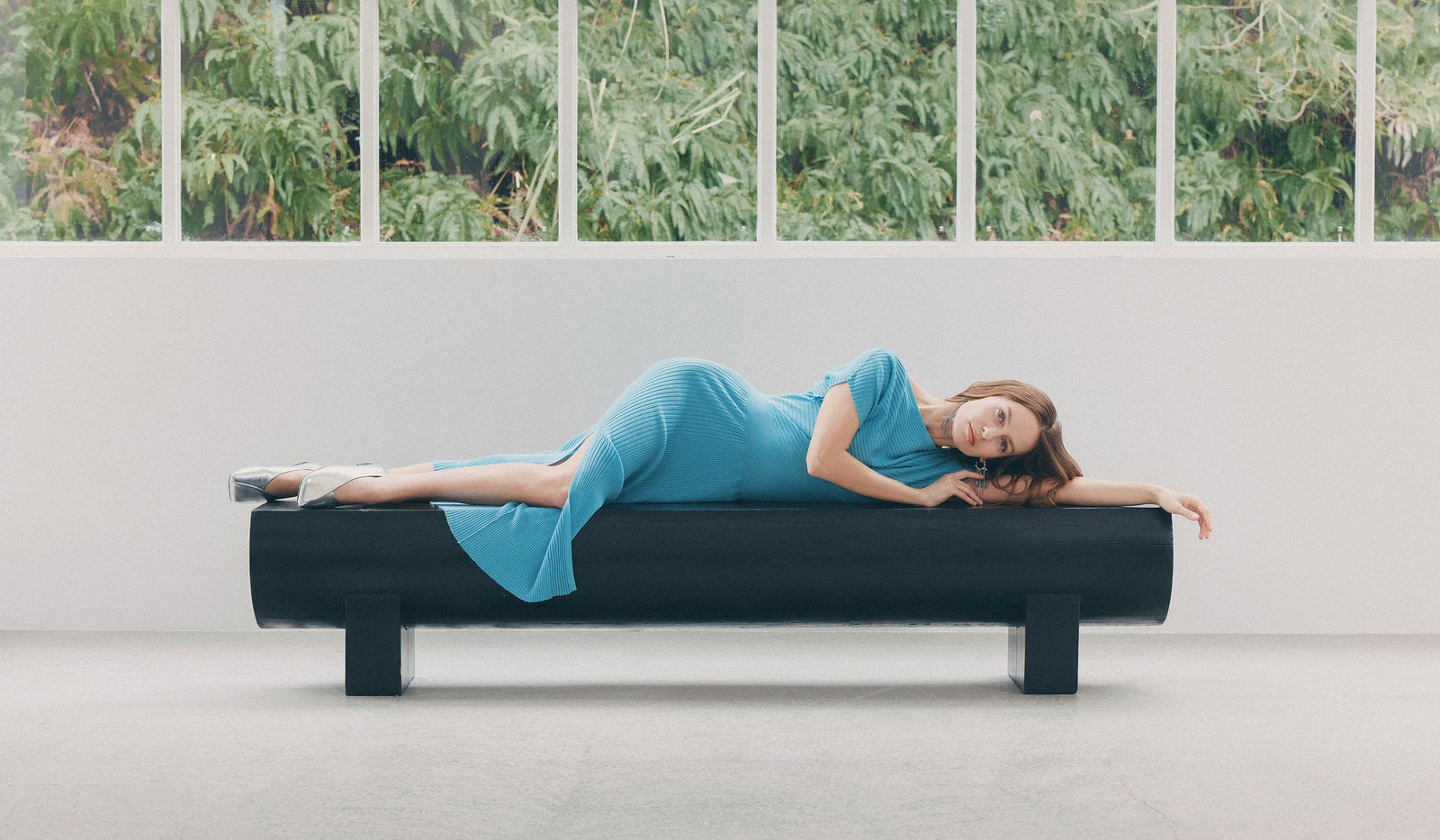 Woman in a blue dress lying on a black bench with a green leafy background.  desktop