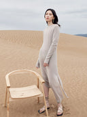 Woman in a long light gray dress standing in a desert with a wooden chair. mobile