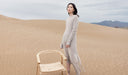 Woman in a long light gray dress standing in a desert with a wooden chair nearby. desktop