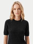 A blonde woman wearing a short-sleeve black cable-knit crewneck sweater, photographed against a clean light gray studio background.