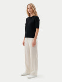 A woman modeling a black short-sleeve ribbed knit top and off-white wide-leg trousers, paired with white sneakers, against a light gray studio background.