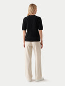 A woman shown from the back wearing a black short-sleeve ribbed knit top and cream wide-leg trousers, standing against a light gray studio background.