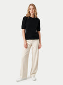 A woman modeling a black short-sleeve ribbed knit top with cream wide-leg trousers and white sneakers, standing against a light gray studio background.