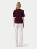 A woman viewed from the back wearing a short-sleeve ribbed knit top in deep burgundy and white wide-leg trousers, set against a plain light gray studio background.