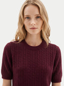 A woman with blonde hair wearing a short-sleeve crewneck cable-knit sweater in burgundy, photographed against a plain light gray background.