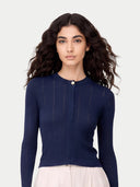 A woman with long, wavy dark brown hair wearing a navy blue ribbed knit cardigan with a single button closure, paired with a light cream skirt. She is photographed against a solid, light gray background. #color_dark-indigo