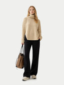 Woman wearing a beige turtleneck sweater and black pants, holding a brown bag on a white background #color_beige
