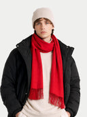 A young man wearing a bright red knit scarf with fringe, a cream beanie, a cream sweater, and a black puffer jacket, photographed against a solid white background.