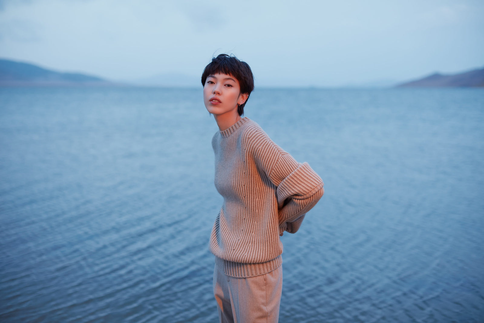 A woman in a peach CashmeReborn sweater standing at the edge of a lake, hills behind