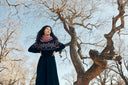 A woman in a dark cashmere dress and pink scarf standing beneath bare trees