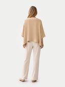 back view of woman wearing a beige poncho and white pants on a white background #color_beige