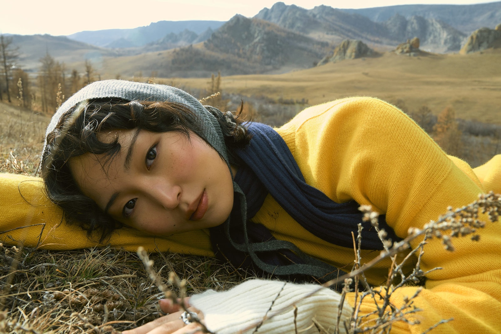 A woman in a yellow cashmere sweater and headscarf lying in autumn grass, hills behind