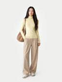 Woman wearing a light yellow turtleneck and beige pants, holding a brown bag on a white background #color_wax-yellow