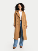Woman wearing a long tan, double breasted coat over a white shirt and blue jeans on a white background  #color_almond