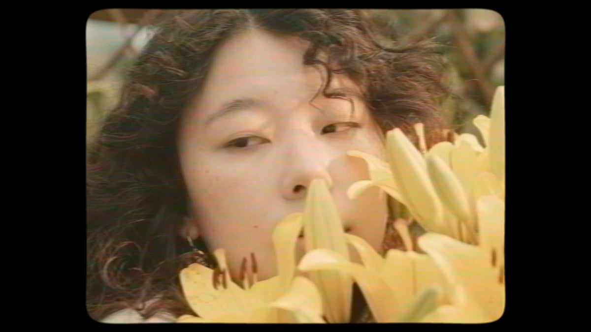 Woman with dark curly hair behind yellow lilies, a still from the campaign film