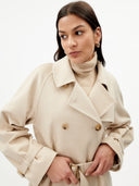 A close-up shot of a person wearing a cream-colored, double-breasted trench coat with large brown buttons and a matching belt. Underneath, they are wearing a tonal beige turtleneck sweater. The person is looking off-camera and wearing small gold hoop earrings. #color_beige